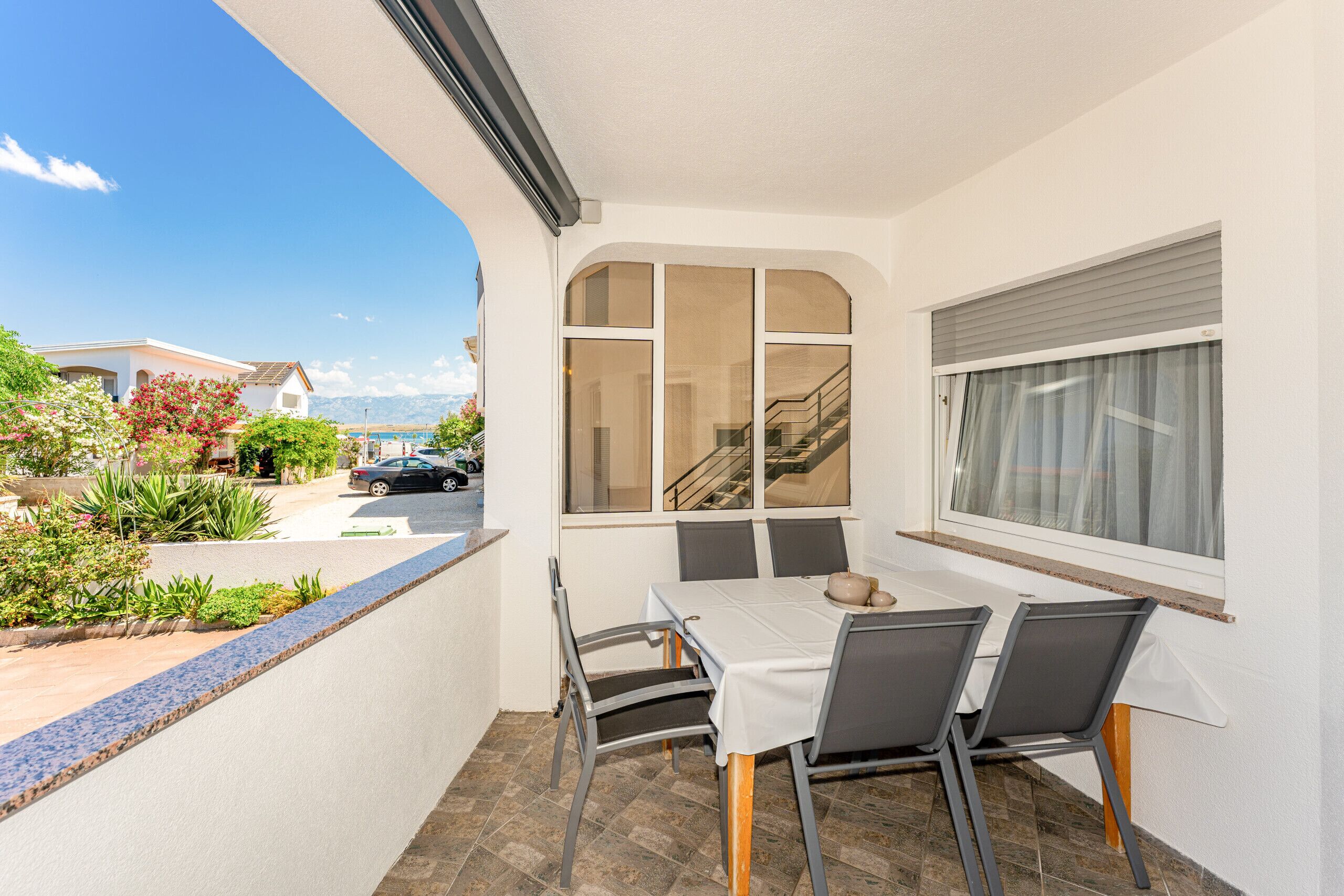 A balcony with a table and chairs, a view of a street and houses, and a window with blinds.