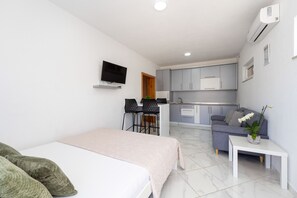 A modern hotel room with a bed, a small table, a sofa, a TV, a kitchenette, and a bathroom.