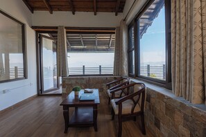 Una habitación con piso de madera, pared de piedra, una mesa con una computadora portátil y una planta, y dos sillas. Hay ventanas grandes con cortinas y vistas al exterior.