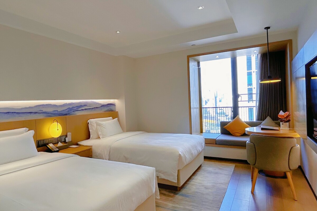 A hotel room with two beds, a view of the city, a small table with a lamp, and a large window.