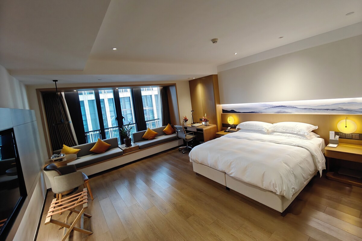 A modern hotel room with a large bed, a desk, a sofa with cushions, a TV, and a large window with a view of buildings.