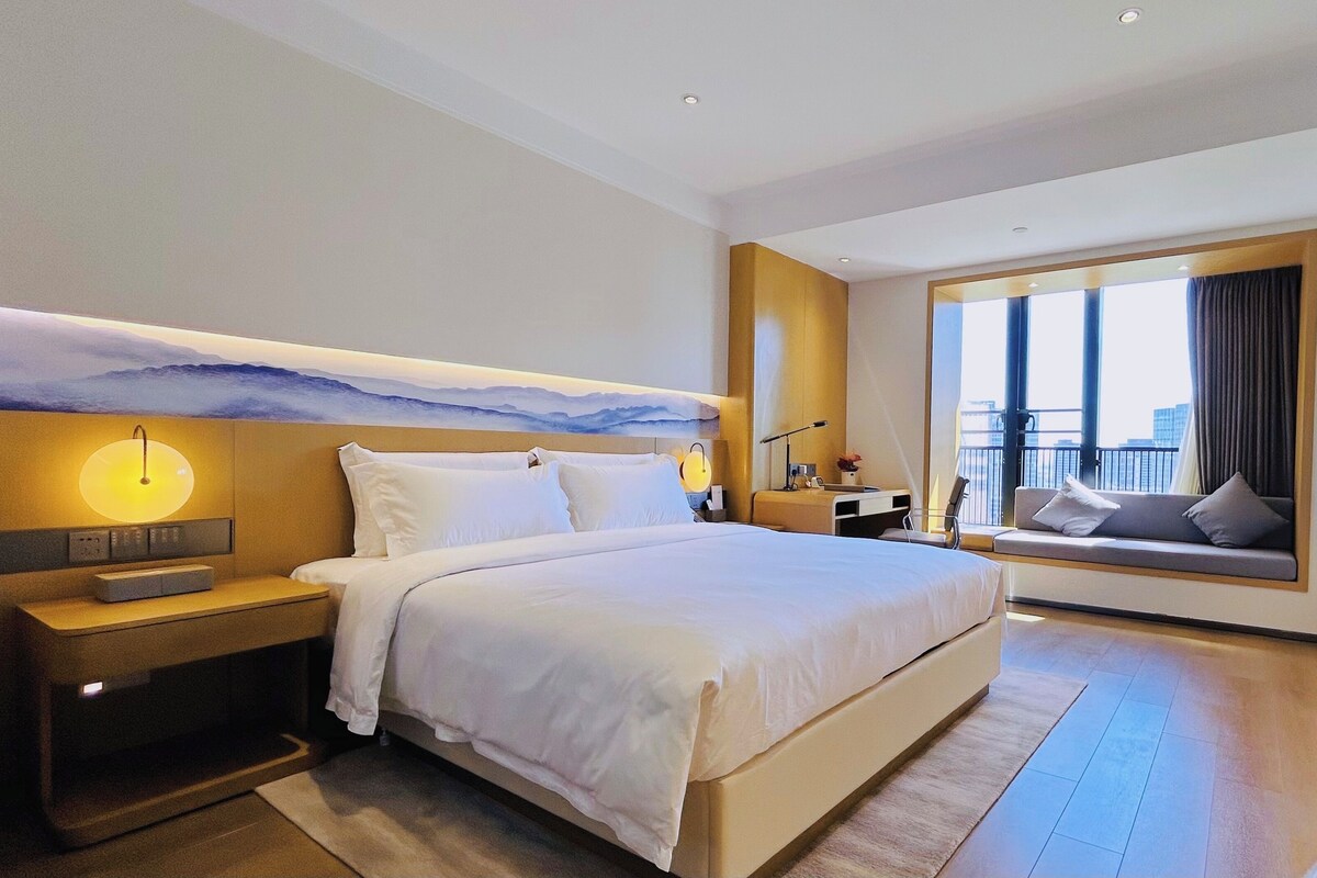 A modern hotel room with a large bed, bedside tables, a wall-mounted artwork, a seating area with a view, and a large window.