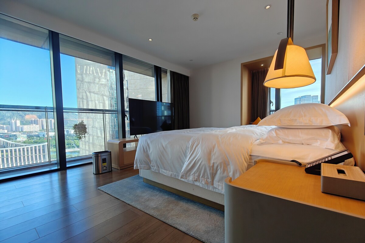 A modern hotel room with a large bed, a TV, and a view of the cityscape through floor-to-ceiling windows.