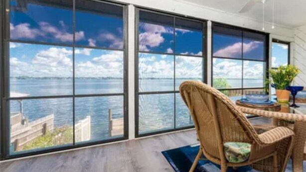 A room with large windows offering a view of the sea and a dock. There is a wicker chair and a table with a blue rug.