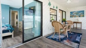A modern room with a glass sliding door leading to a balcony, a dining area with a table and chairs, and a bed with a blue patterned bedspread.