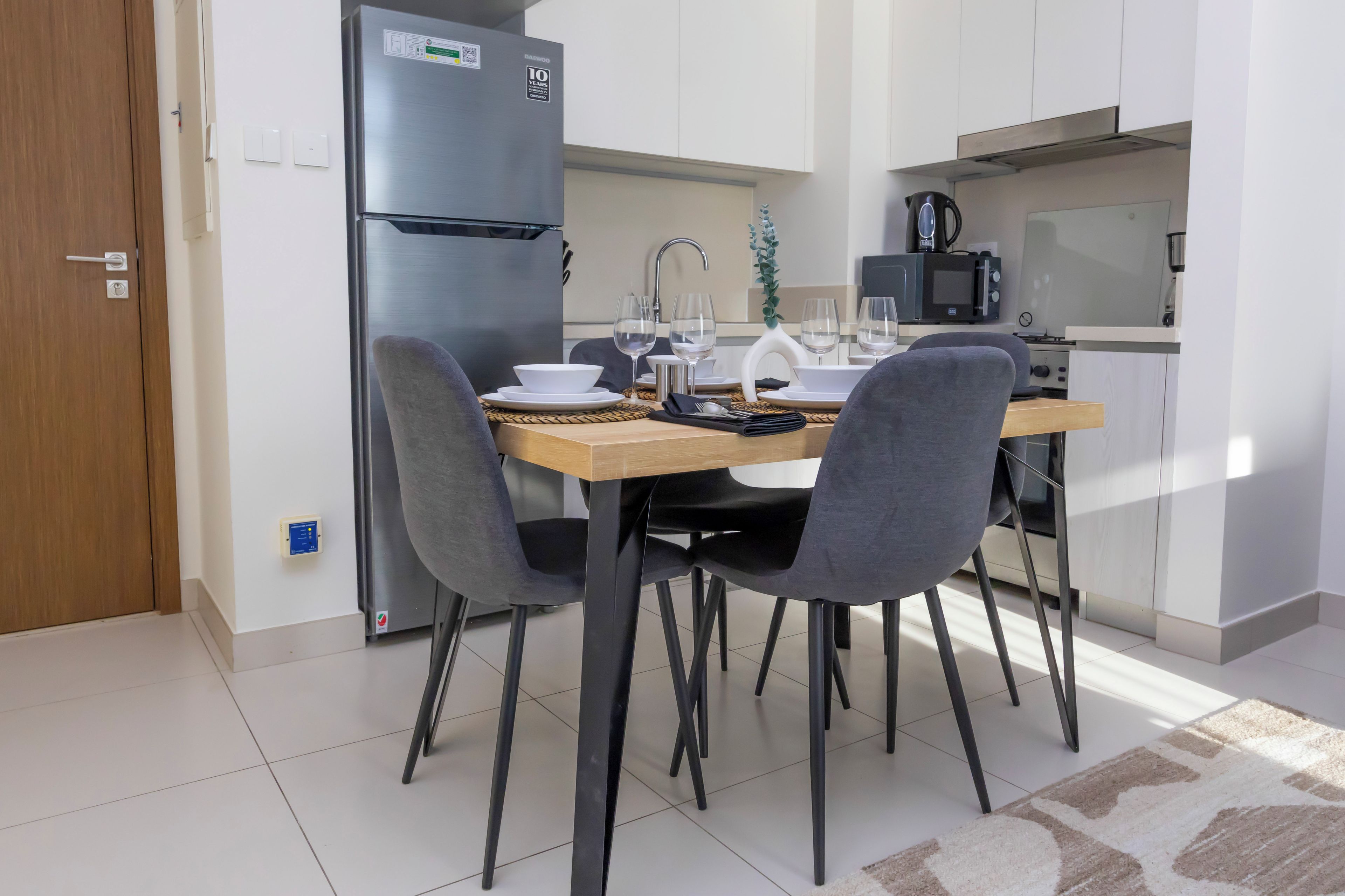 Une cuisine moderne équipée d’un réfrigérateur en acier inoxydable, d’un four à micro-ondes et d’une bouilloire. On y trouve quatre chaises disposées autour d’une table en bois, sur laquelle est posé un set de table.
