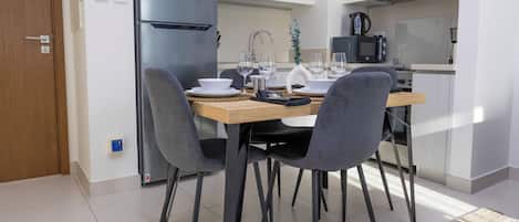 A modern kitchen with a stainless steel refrigerator, a microwave, and a kettle. There are four chairs around a wooden table with a placemat.