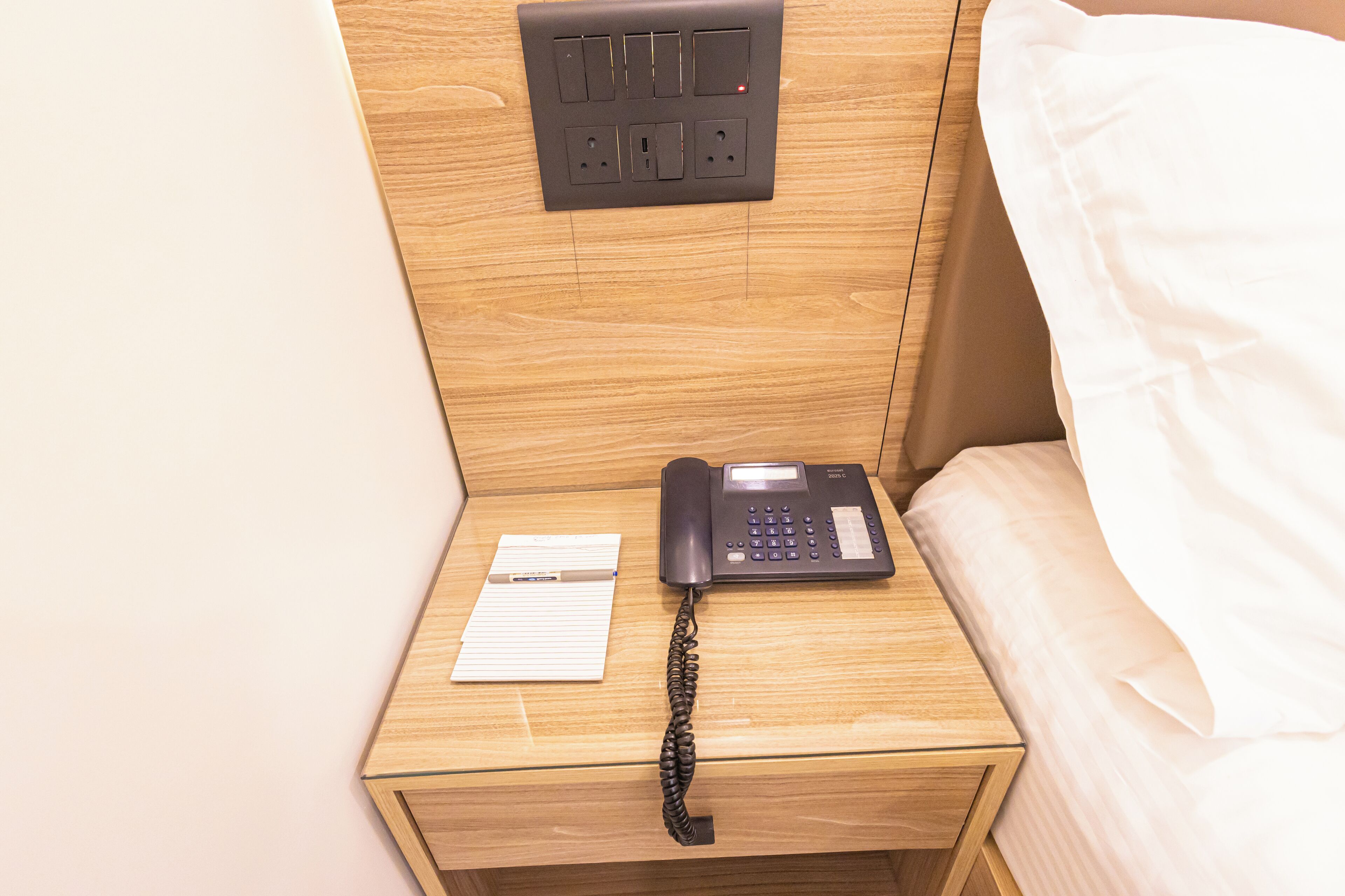 A wooden bedside table with a black telephone, a notepad, and a power outlet.