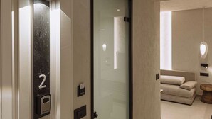 Un pasillo moderno con puerta de cristal, un letrero con el número '2', un lector de tarjetas de acceso y una zona de estar minimalista con un sofá y una mesa redonda de madera.