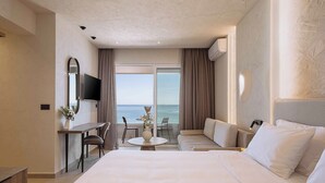Habitación de hotel con una cama grande, un escritorio con silla, una mesita, un sofá, un televisor montado en la pared y vista al mar a través de la ventana.