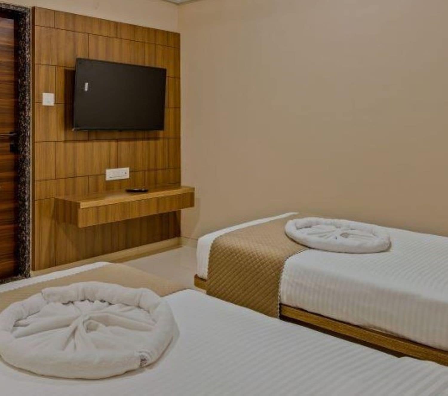 A hotel room with two beds, a TV mounted on a wooden panel wall, and a small shelf below the TV.