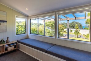 A room with a built-in bench along a window, a small shelf with books and a vase, and a view of greenery and a building across the street. - Cabbage Tree Cottage - Coastal Comfort by the Bay (Waiheke Island)