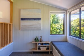 A room with a view of a beach, a bench, a shelf with books and a vase, and a painting on the wall. - Cabbage Tree Cottage - Coastal Comfort by the Bay (Waiheke Island)