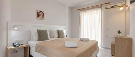 A modern hotel room with a large bed, bedside table with a phone, a small desk, a mirror, and a window with curtains.