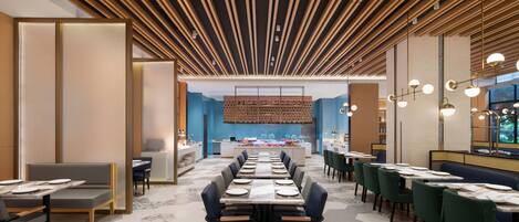 A modern dining area with a long table, multiple chairs, and a kitchen in the background.