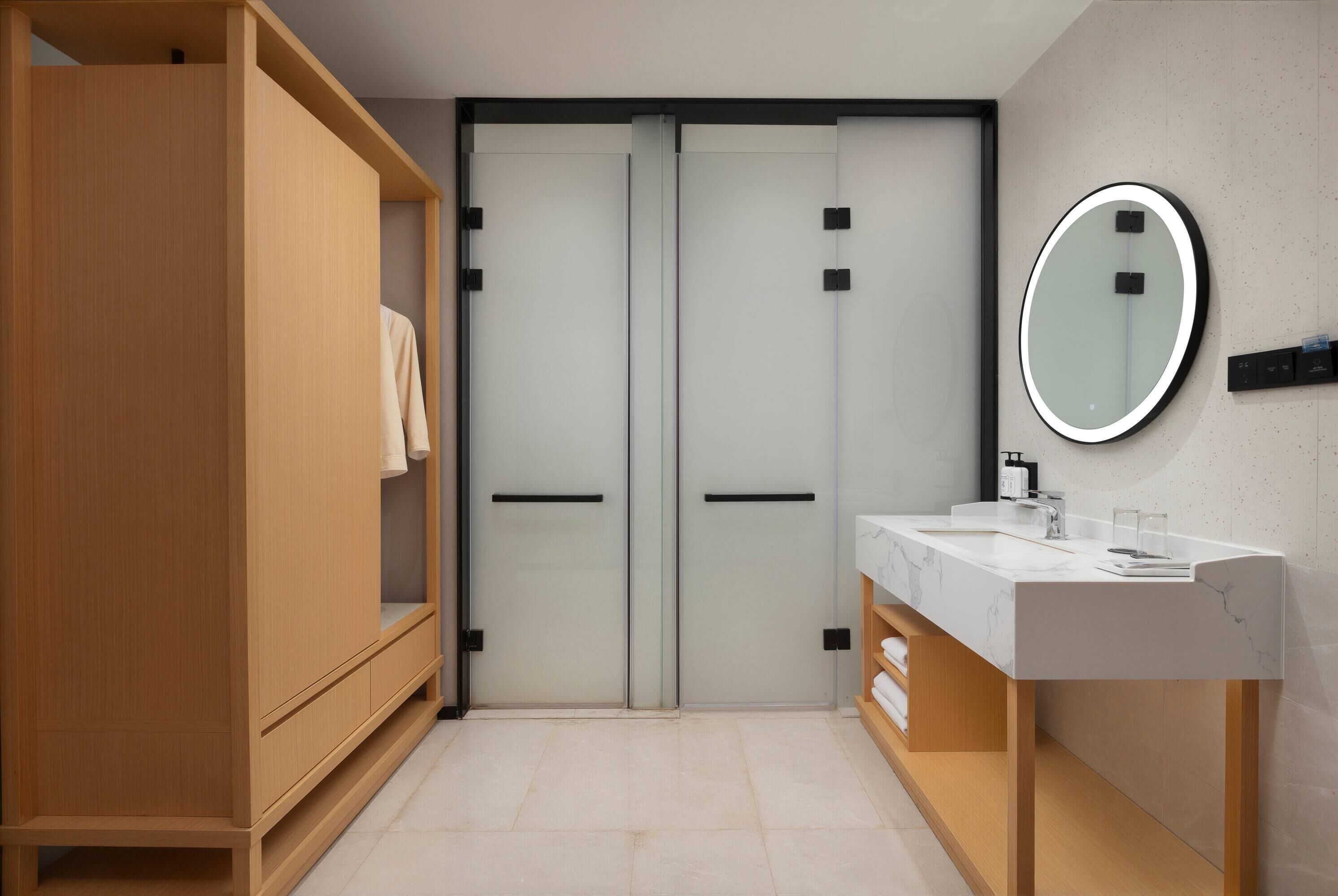 Un baño moderno con un armario de madera, un closet con puerta de vidrio, un tocador blanco con encimera de mármol y un espejo redondo.