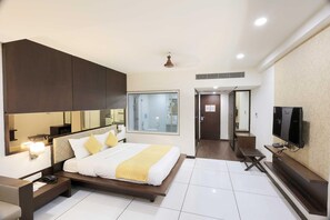 Una habitación de hotel moderna con una cama grande, mesita de noche, televisor y un ventanal amplio.