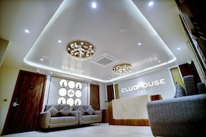 Un vestíbulo moderno con un mostrador de recepción identificado como "CLUBHOUSE", una puerta de madera, un sofá color beige y un sillón gris.