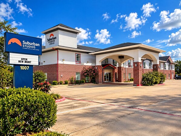 A Suburban Studios hotel with a brick facade, a sign indicating an open new management, and a parking lot.