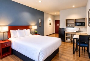 A hotel room with a bed, bedside tables, a desk, a chair, a microwave, and a refrigerator.