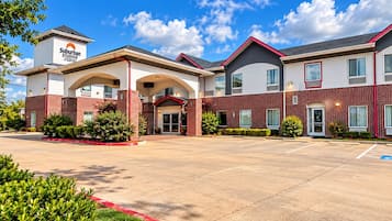 Un hotel Suburban Studios con una entrada central, varios espacios de estacionamiento y un cielo despejado.