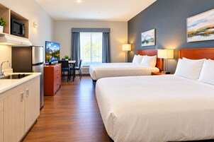 A hotel room with two beds, a kitchenette, a desk with a chair, and a television.
