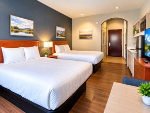 A hotel room with two beds, a desk, a TV, and a kitchenette.