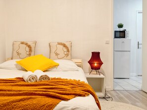 Uma cama bem arrumada com um travesseiro amarelo, uma luminária vermelha e uma geladeira branca ao fundo.