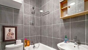 Kylpyhuoneessa on valkoinen kylpyamme, valkoinen pesuallas ja wc-istuin. Pesualtaalla on hygieniatuotteita ja seinällä on kehystetty kuva.