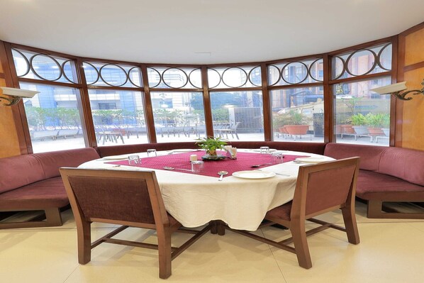 A dining area with a round table set for four, surrounded by cushioned seating. Large windows with decorative frames offer a view of the outside.