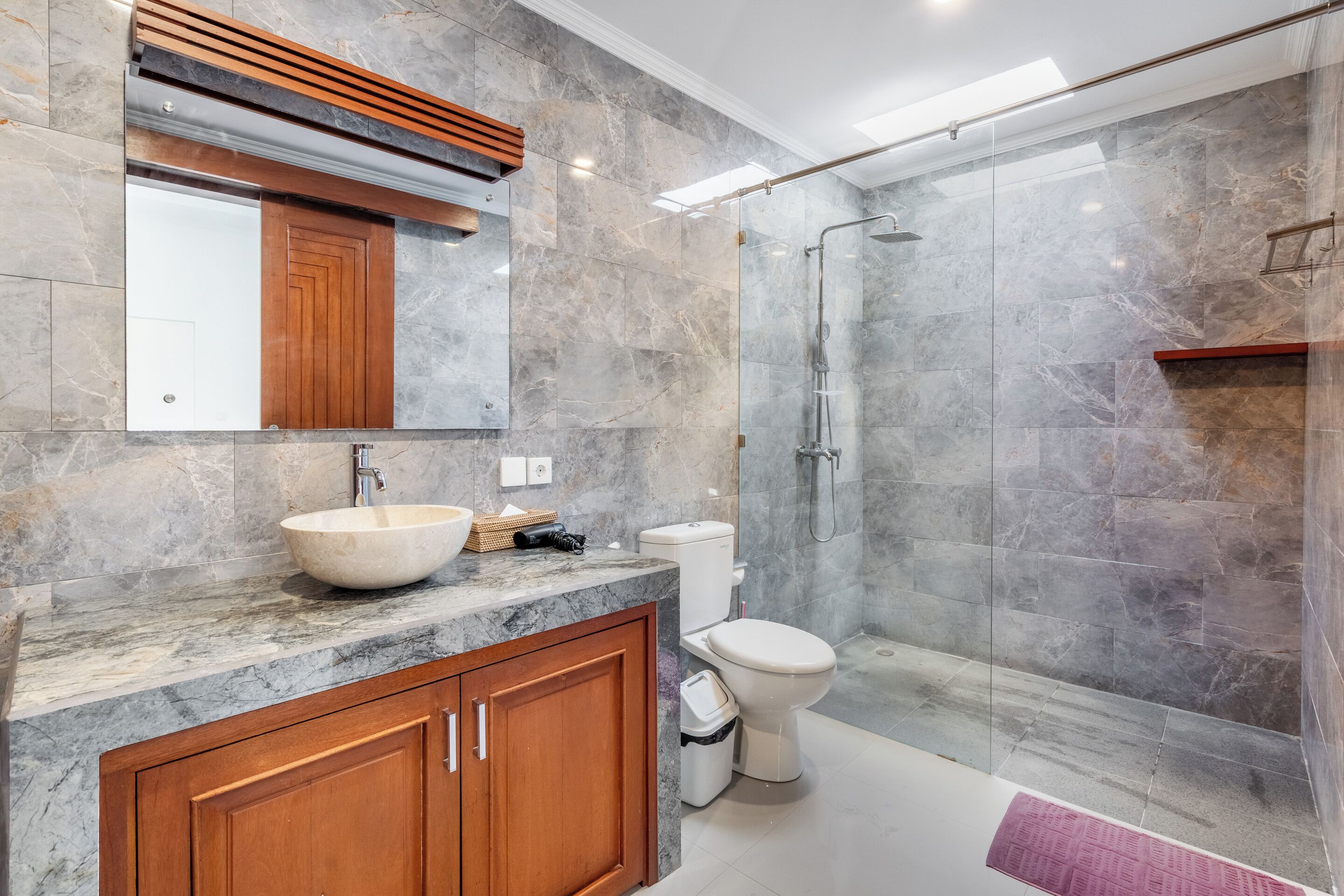 Un baño con lavamanos de mármol, armarios de madera, inodoro y cabina de ducha con paredes de vidrio.