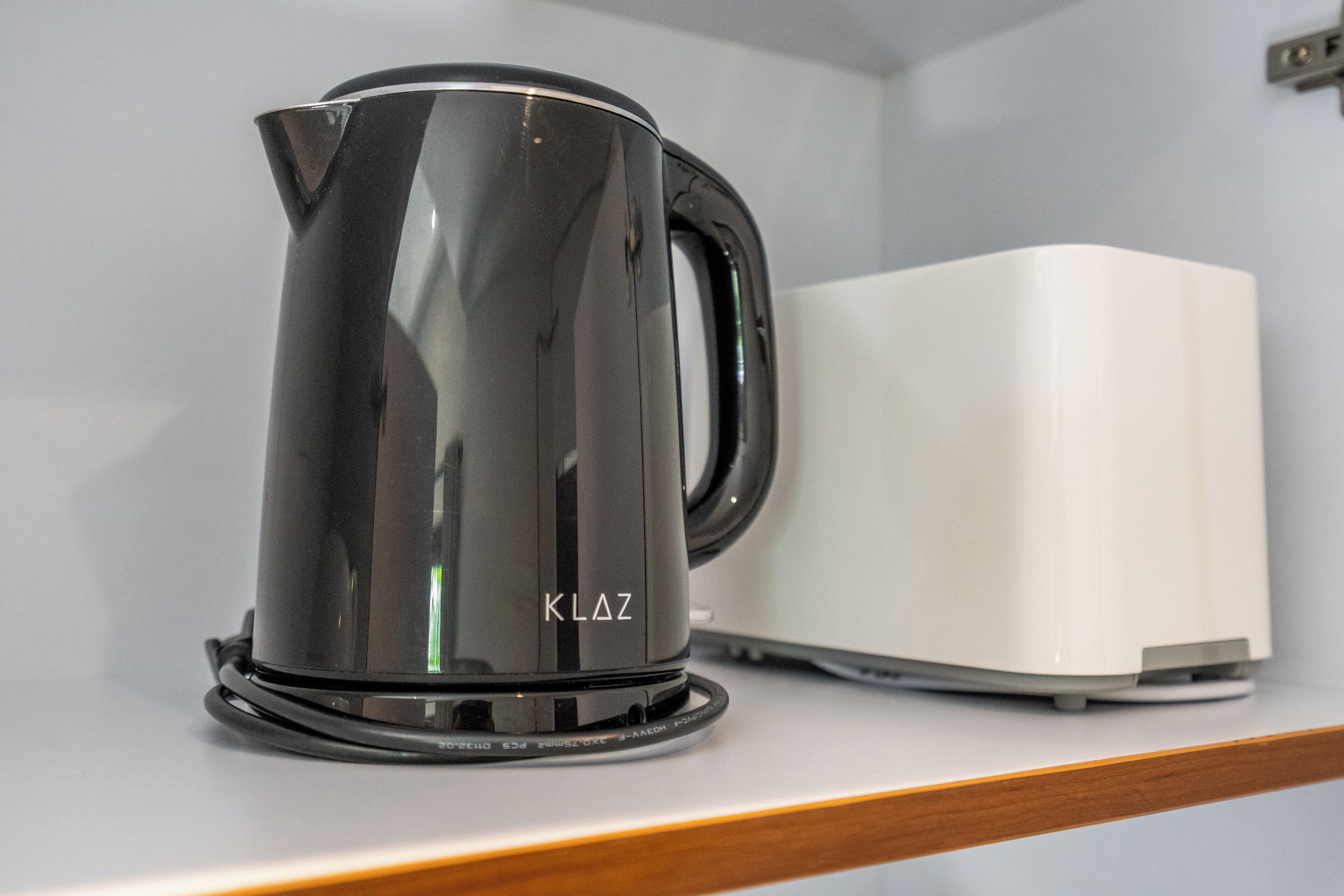 Una tetera negra con la marca 'KLaz' sobre una encimera blanca, junto a un electrodoméstico blanco.