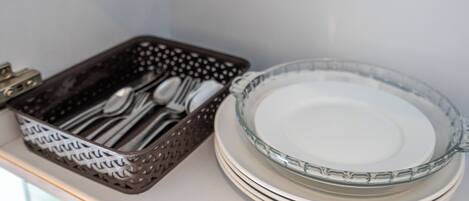 A stack of white plates and a glass bowl on a white surface, with a brown basket containing silverware.