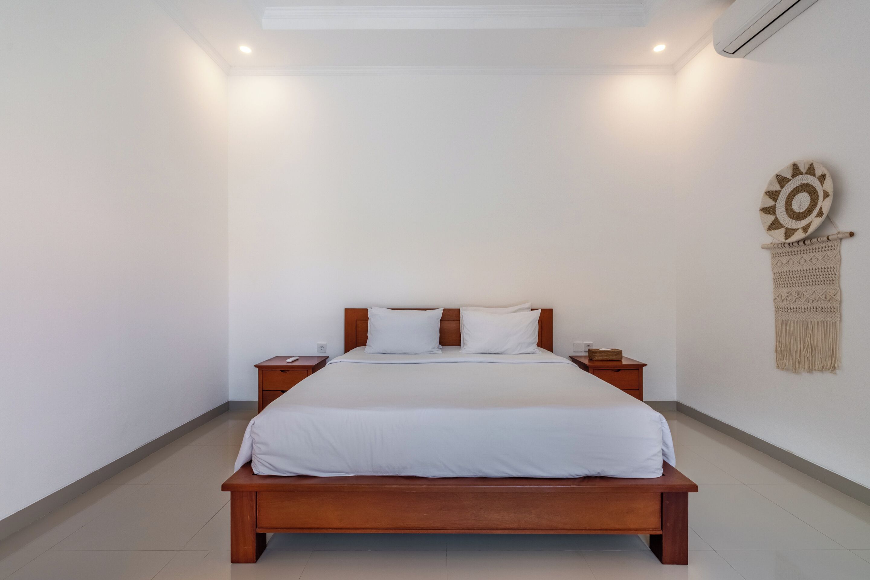 Ein Schlafzimmer mit einem hölzernen Bett, weißer Bettwäsche, zwei Nachttischen, einer Wanddekoration und einer Klimaanlage.