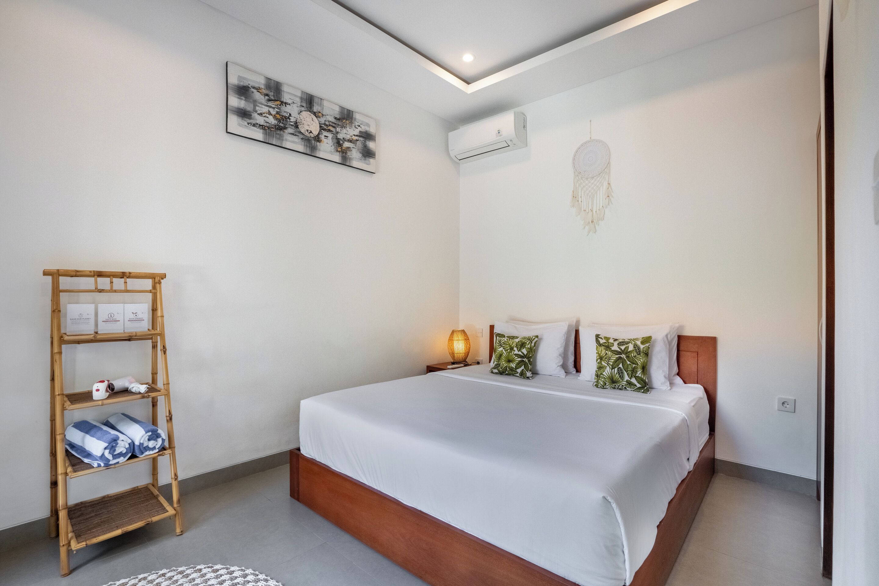Un dormitorio con cama, lámpara de noche, aire acondicionado mural, una obra de arte enmarcada y estante de madera con toallas.