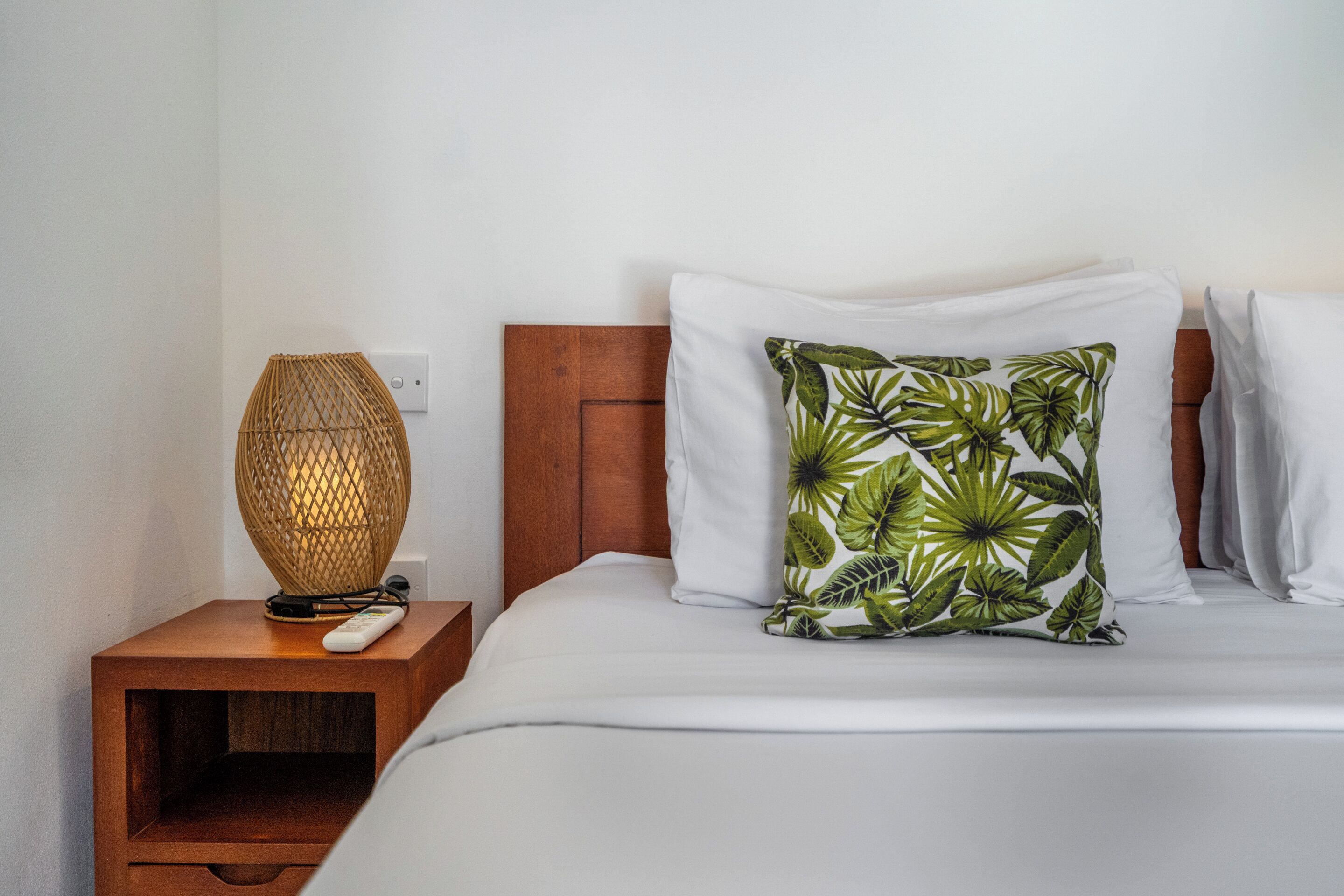 Un lit bien fait, avec une couette blanche, une tête de lit en bois et un seul coussin décoratif orné d’un motif de feuilles tropicales.