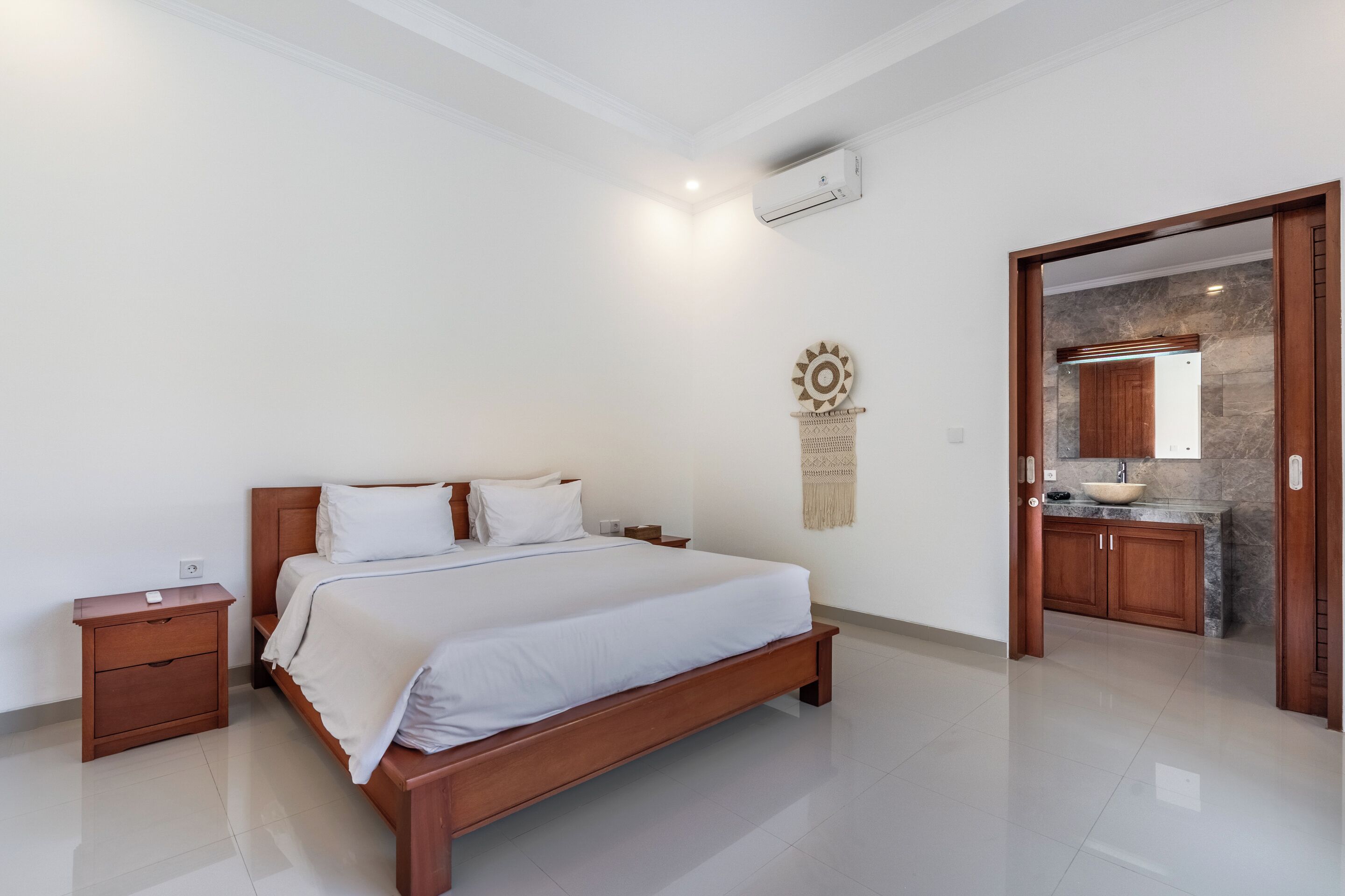 A bedroom with a wooden bed, a nightstand, a wall-mounted air conditioner, and a kitchen area visible through an open door.