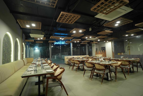 A restaurant with long tables and cushioned seating, modern lighting fixtures, and a view through large windows.