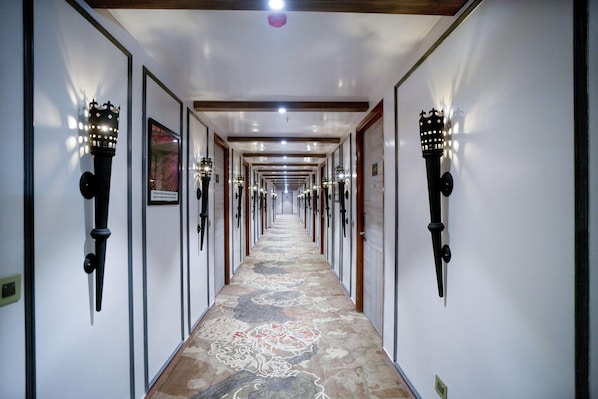 A hotel hallway with a patterned carpet, wall-mounted sconces, and framed pictures on the walls.