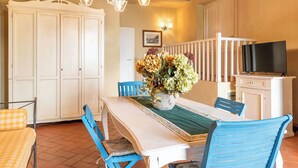 Ein Essbereich mit einem Tisch für vier Personen, blauen Stühlen, einer Blumenvase, einer Chandelüster und einem Fernseher auf einem Schrank.