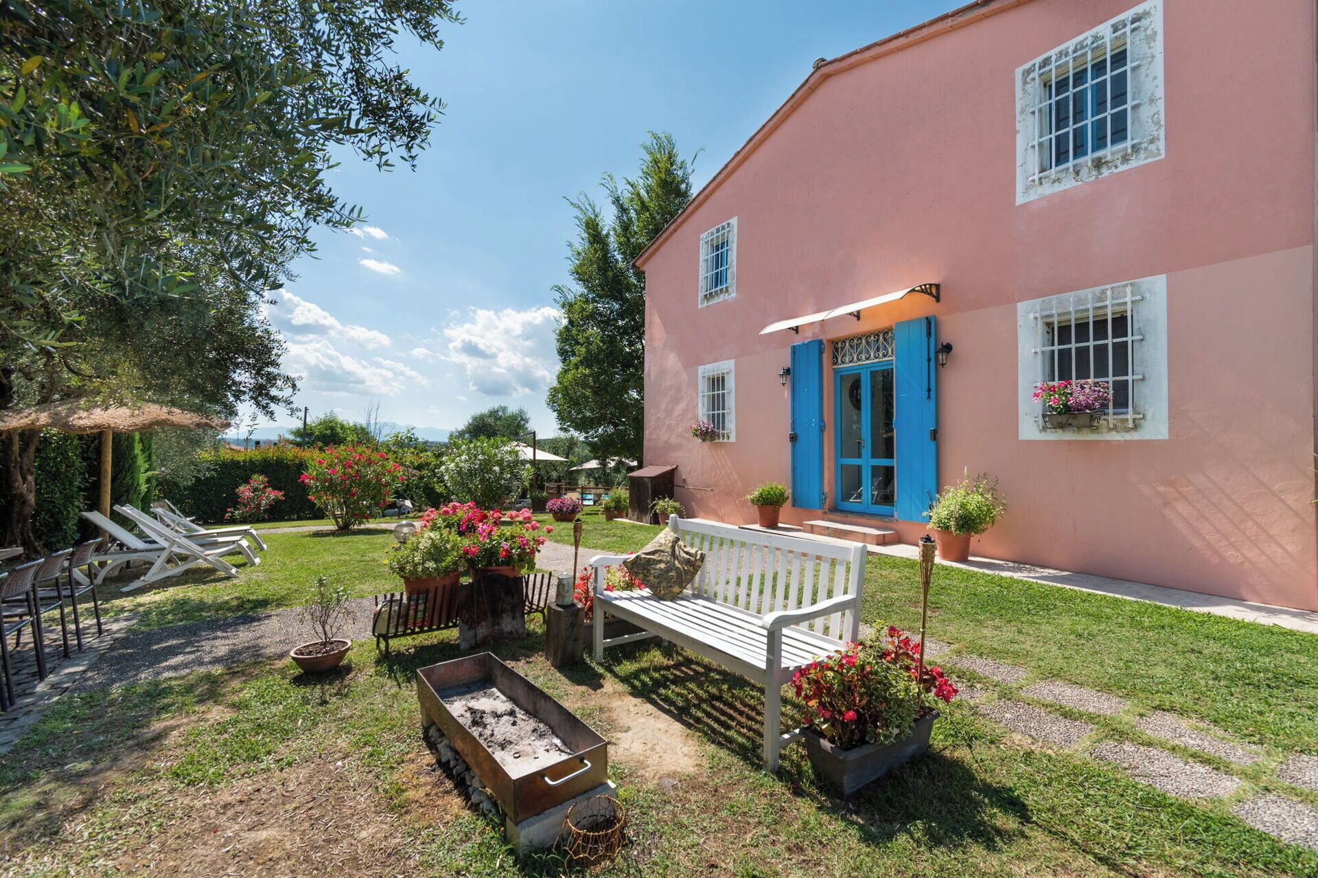 A pink building with a blue door and windows, surrounded by a garden with potted plants and a bench.