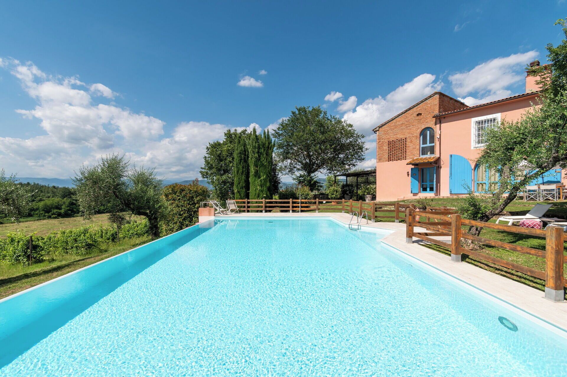 A clear blue swimming pool with a wooden deck and lounge chairs, surrounded by a wooden fence and trees.