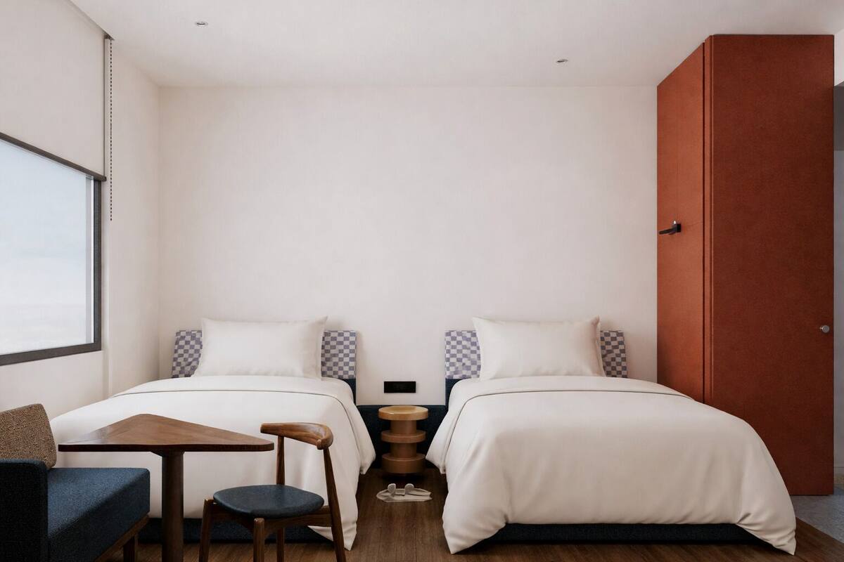A hotel room with two beds, a small table with a chair, a wooden side table, and a large window.