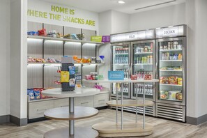 Un bar de aperitivos en un hotel, con estantes llenos de bocadillos y bebidas, una mesa y un letrero que indica la ubicación de los productos.
