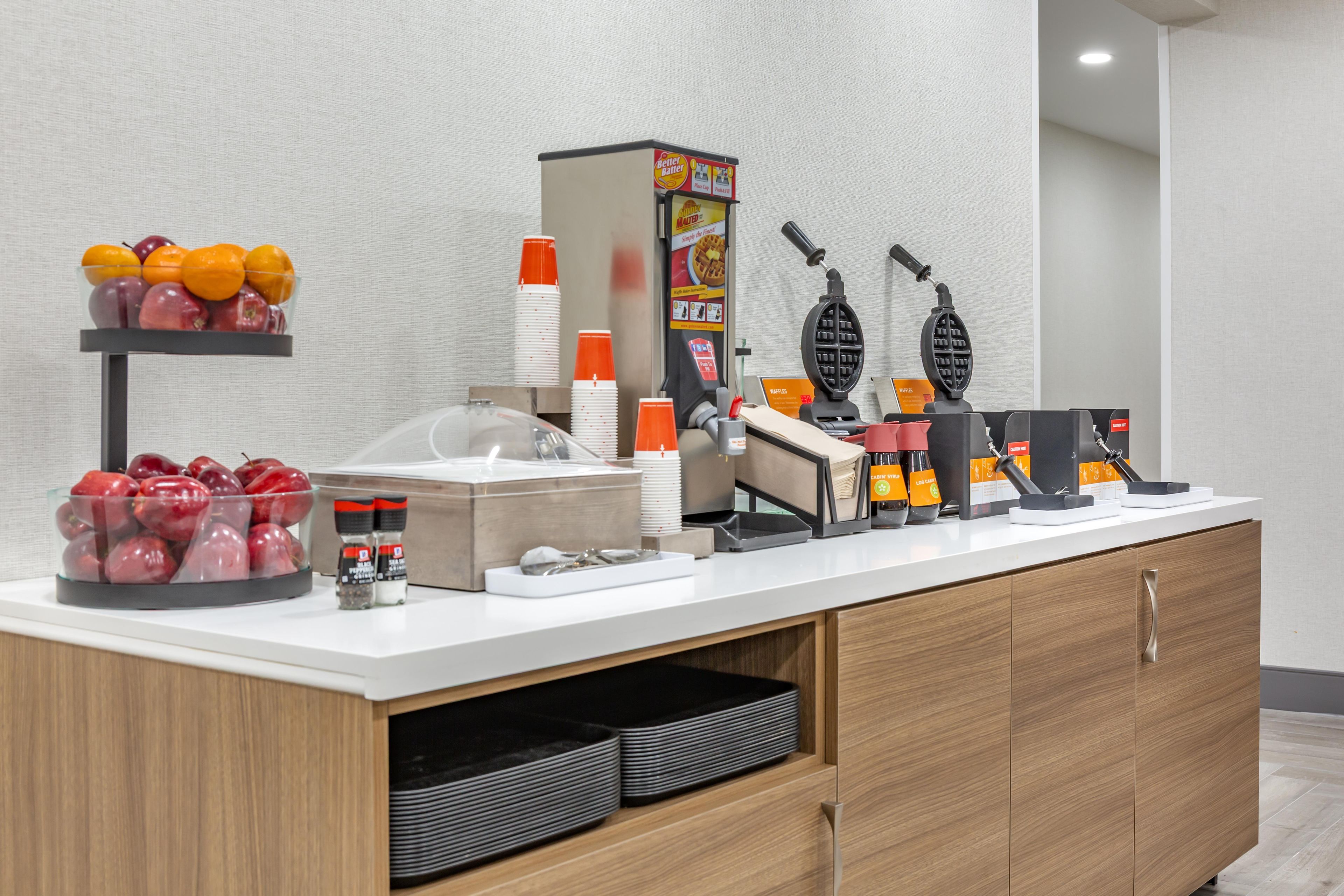 A breakfast bar with fruit, condiments, waffle makers, and disposable cups.
