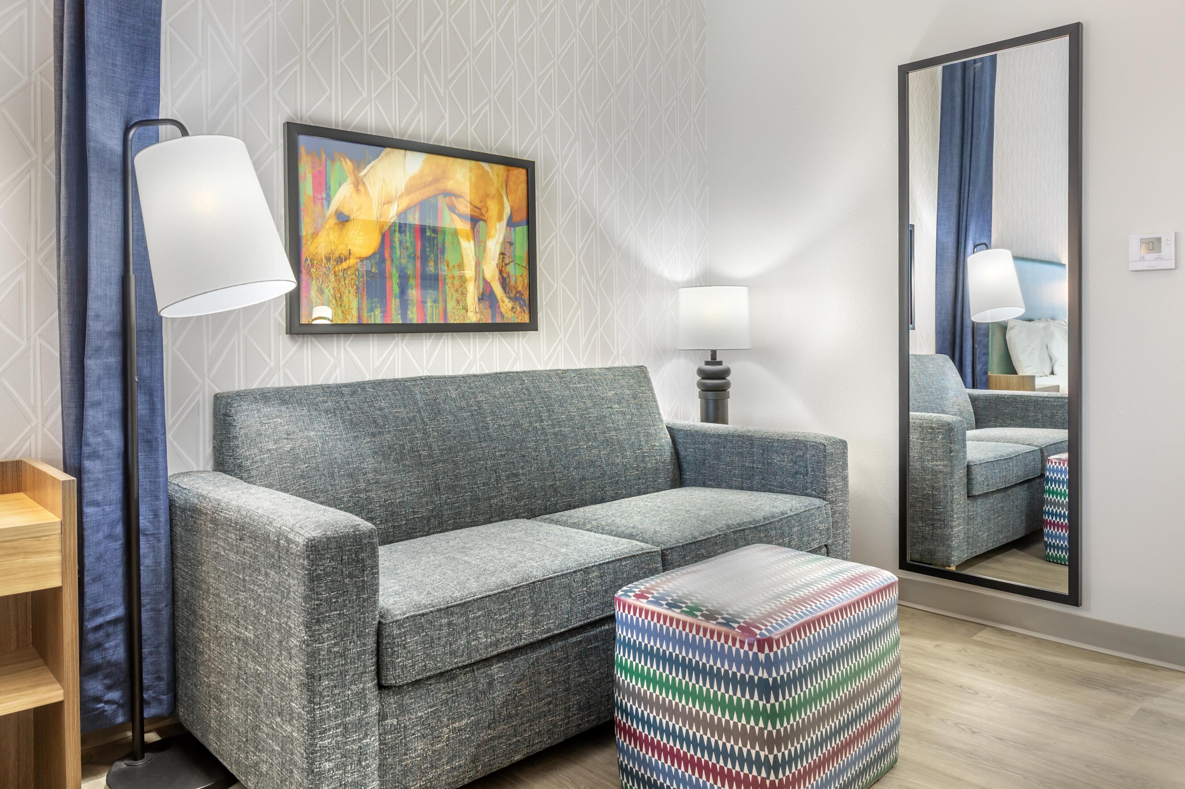 A modern hotel room with a grey sofa, a colorful abstract painting, a side table, a lamp, a mirror, and a patterned ottoman.