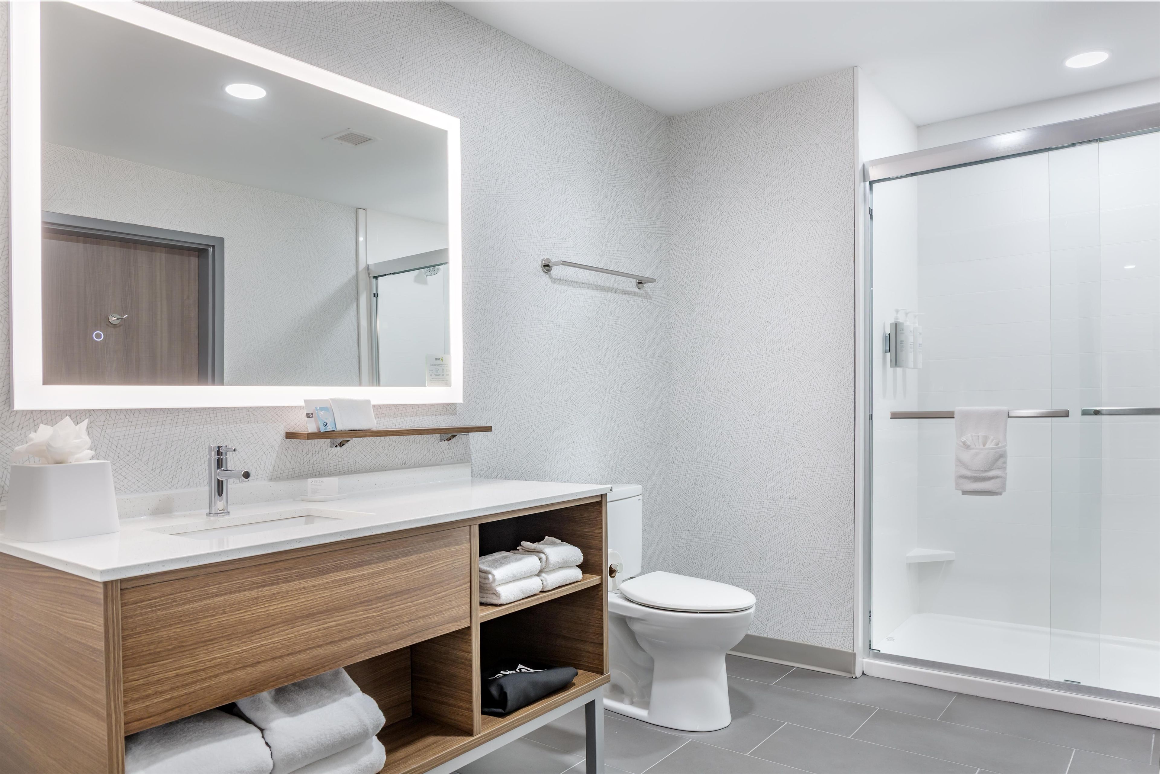 Un baño moderno con un espejo grande, un tocador de madera, un inodoro y una cabina de ducha de vidrio.