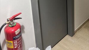 Uma porta cinzenta com puxador prateado, uma parede branca com interruptor de luz e um extintor de incêndio vermelho no chão.