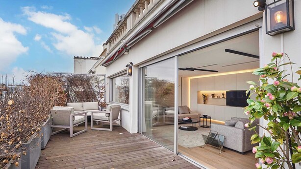 Eine moderne Terrasse im Freien mit Holzdeck, einer Sitzecke mit Sofa und Stühlen sowie einer Glastür zu einem Innenraum mit Fernseher und Dekoration.