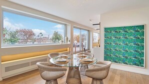 Ein moderner Essbereich mit einem Glastisch, Korbstühlen und einem großen Fenster mit Blick auf Bäume und Gebäude.